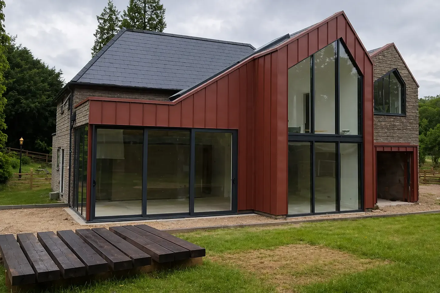 Red zinc standing seam extension with full-height glazing beside a stone house
